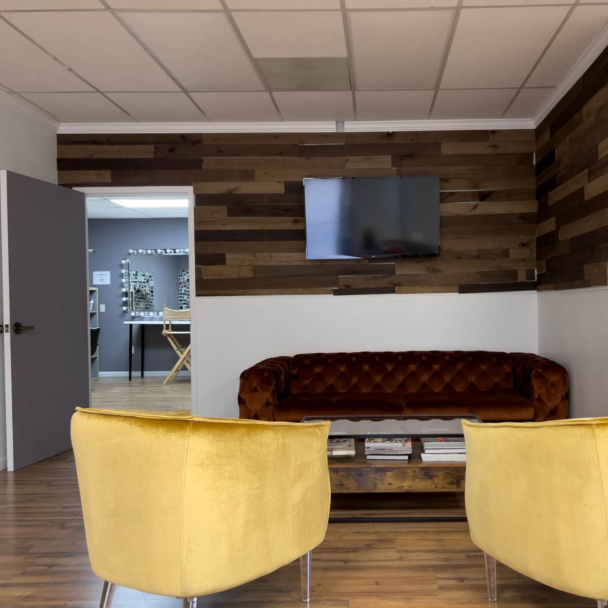 A waiting area with a brown tufted couch, two yellow armchairs, a wooden accent wall, a flat-screen TV, and a fire extinguisher mounted on the wall.