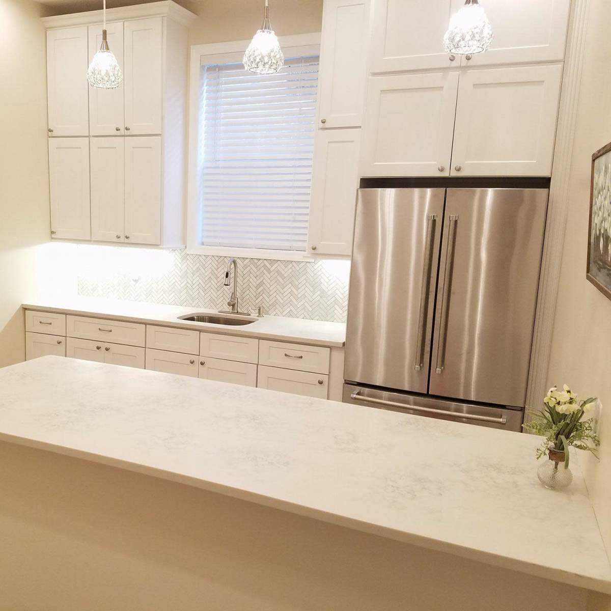 A modern kitchen with white cabinets, a stainless steel refrigerator, and a patterned backsplash, illuminated by pendant lights.