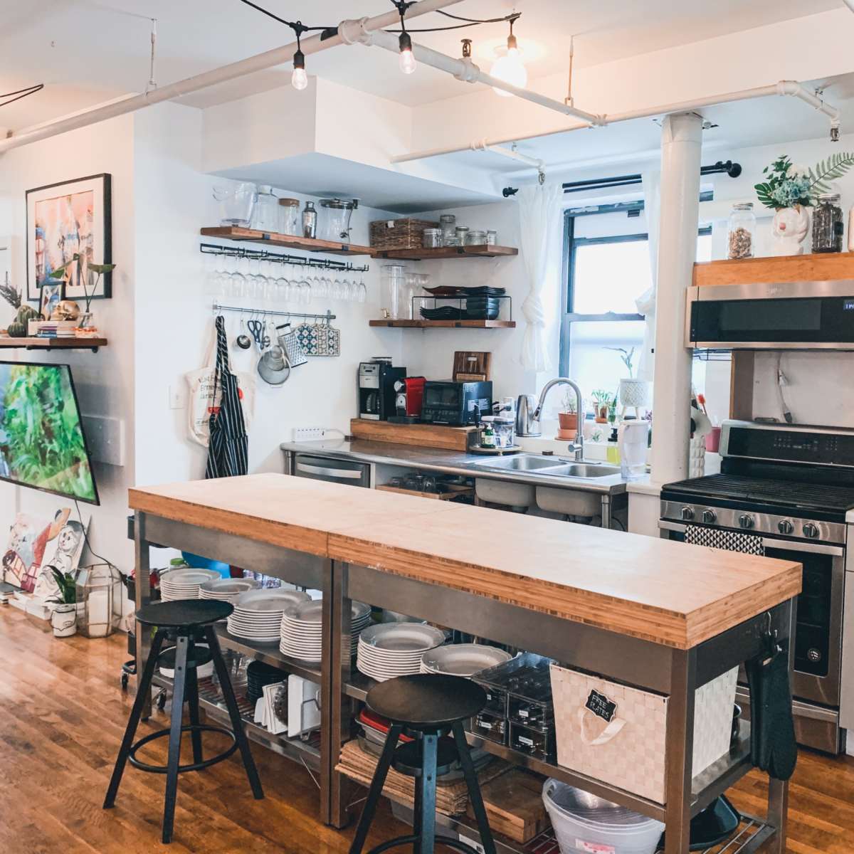 A modern kitchen features an island with a wooden countertop, stools, and shelves stocked with cookware and spices, alongside stainless steel appliances and a sink.