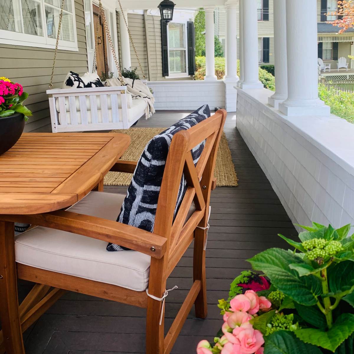 A front porch with a wooden dining table and chair, a swing hanging from the ceiling, and potted plants along the railing.