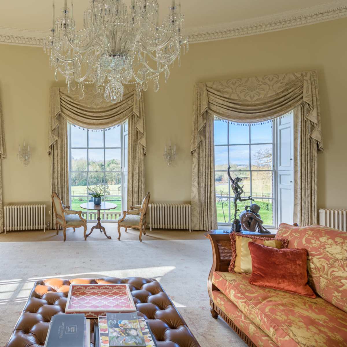 An elegantly furnished room with large windows showcasing a view of a garden and ornate decor including a chandelier and floral-patterned sofas.
