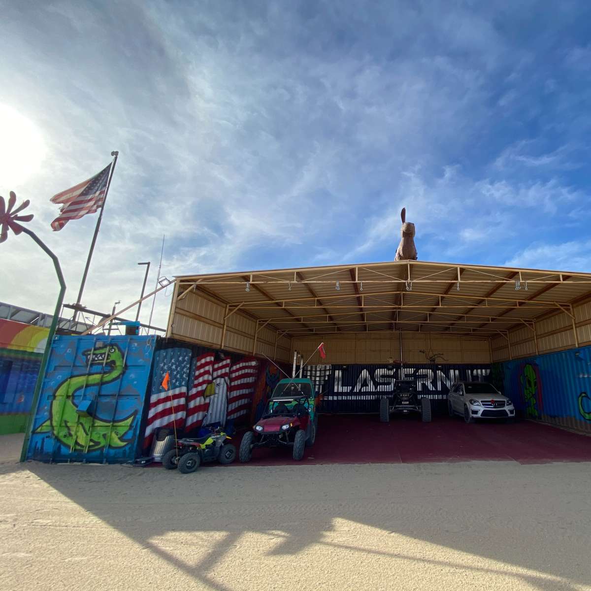 A large, open shed with colorful murals, including dinosaur and flag designs, housing a car and a dune buggy.