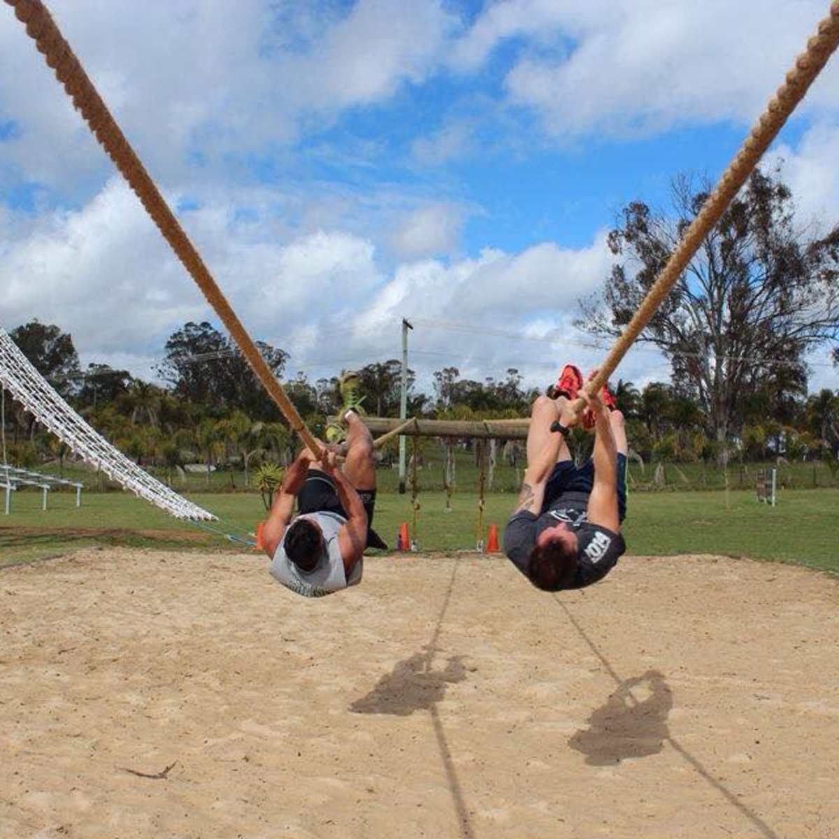 Two men are swinging on ropes above a sandy area in a playground, with equipment and trees in the background.