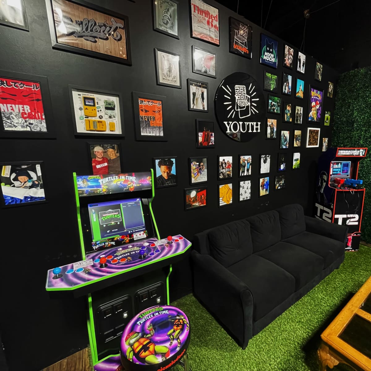 A black wall adorned with framed artwork and posters, featuring a green carpet area with a black couch, a multi-colored arcade machine, and a wooden coffee table.
