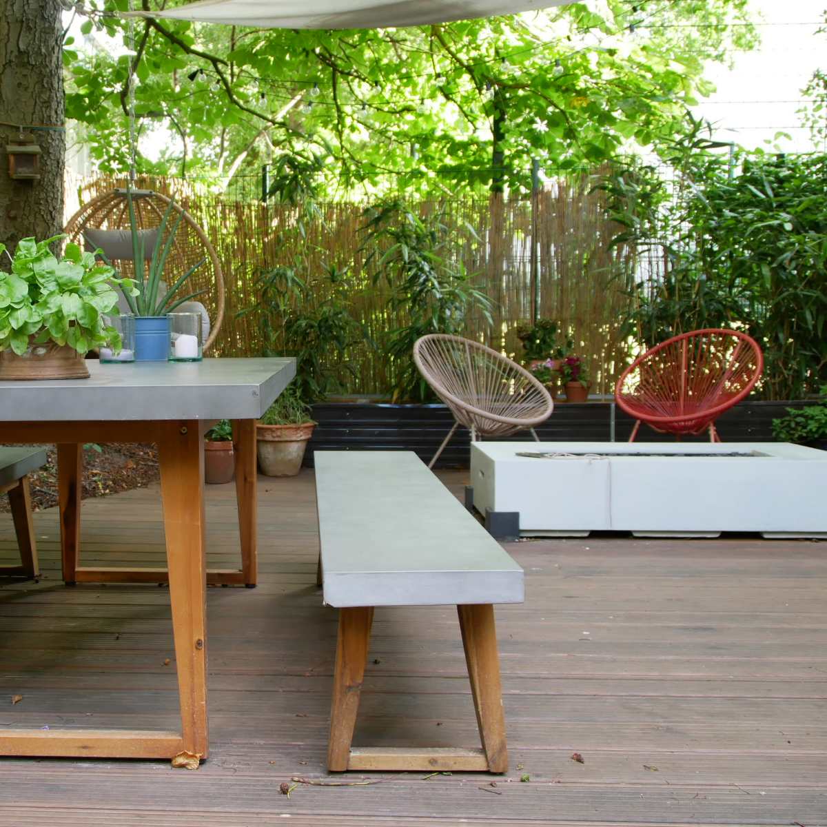 A wooden outdoor dining table with benches is surrounded by greenery in a shaded patio area.
