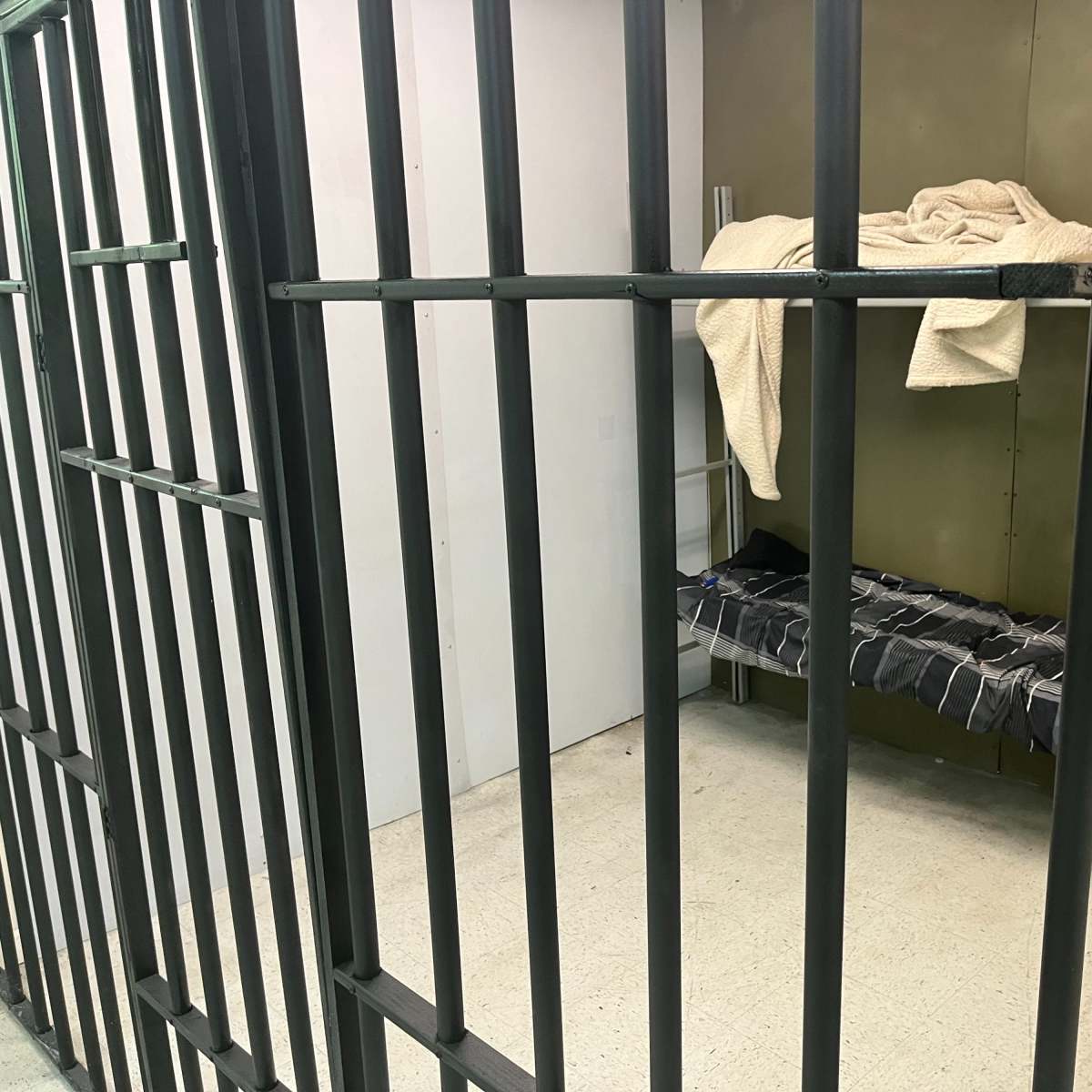 The interior of a prison cell with metal bars, a bed covered with a blanket, and a plain wall.
