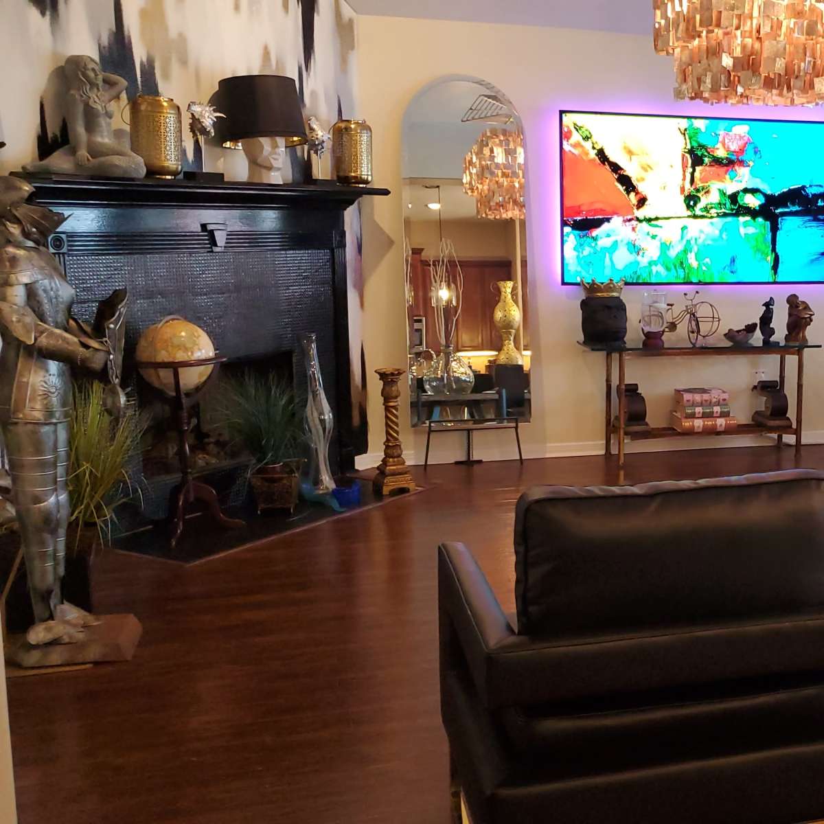 The room features a modern living area with two black chairs facing a glass coffee table, a large television mounted on the wall, and various decorative elements including a fireplace and a large vase with branches.