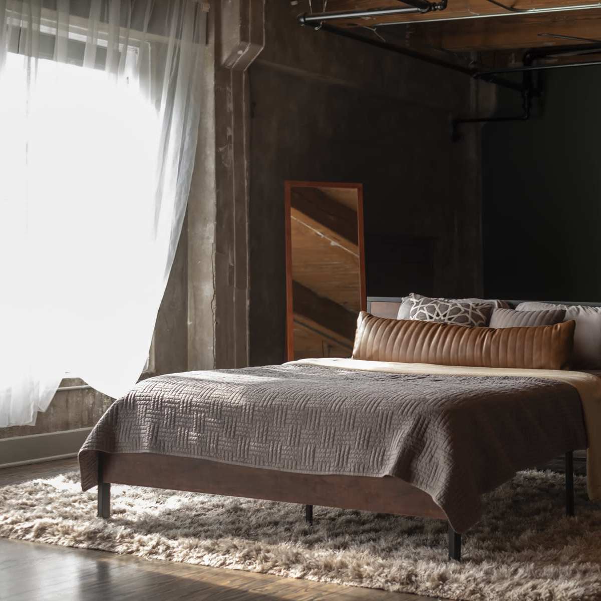 A modern bedroom features a bed with a textured blanket, decorative pillows, and a soft area rug, complemented by large windows and minimalist furniture.