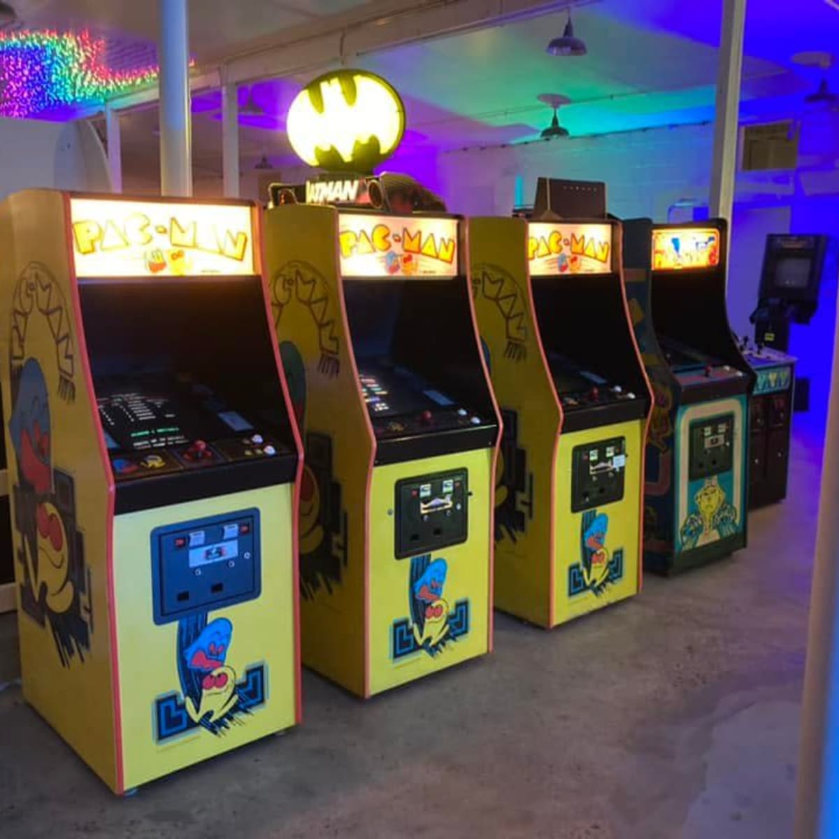 A row of vintage arcade game machines, including Pac-Man, set in a brightly lit arcade with colorful ambient lighting.
