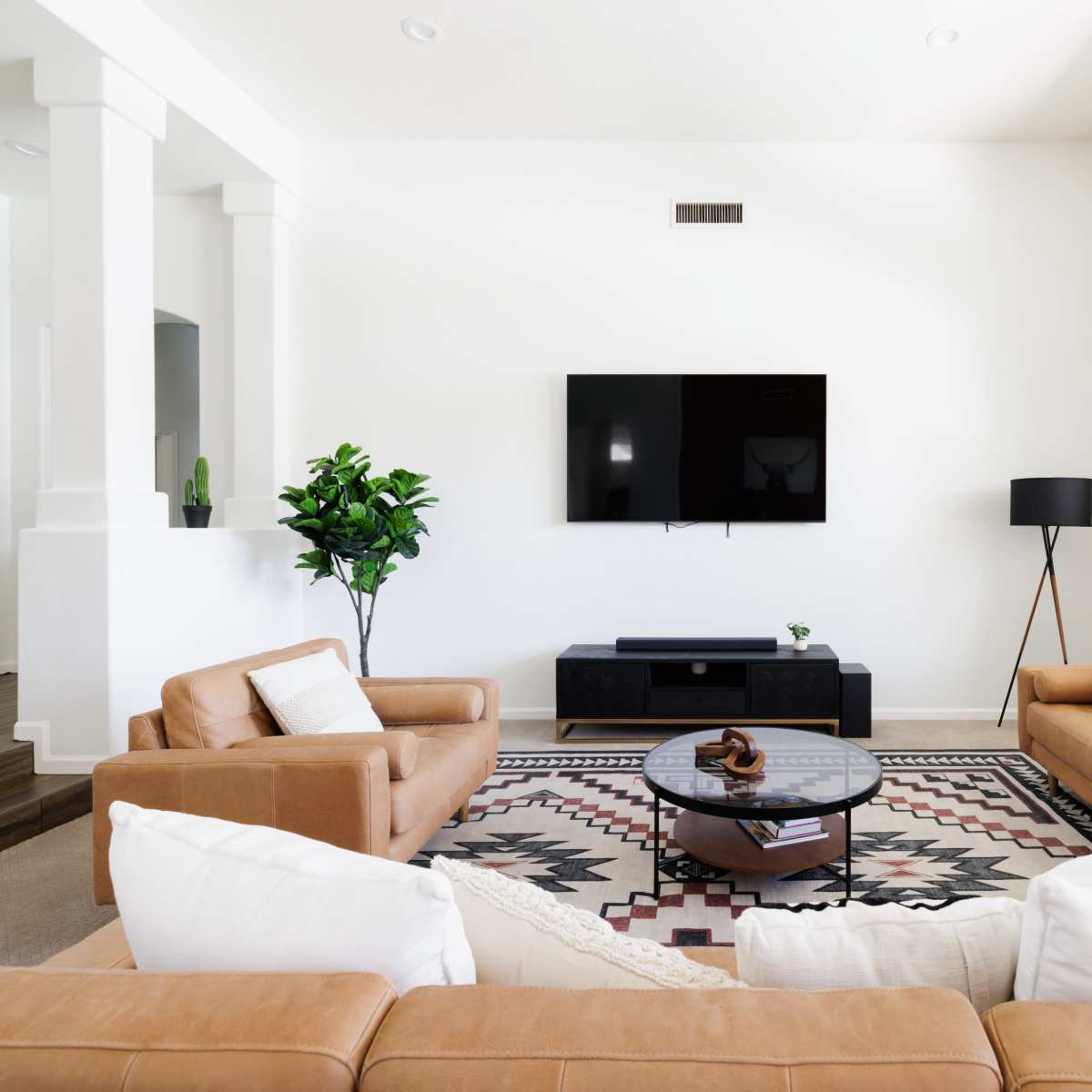A modern living room features two tan leather sofas, a circular coffee table, and a flat-screen TV mounted on a white wall, with large windows allowing natural light.