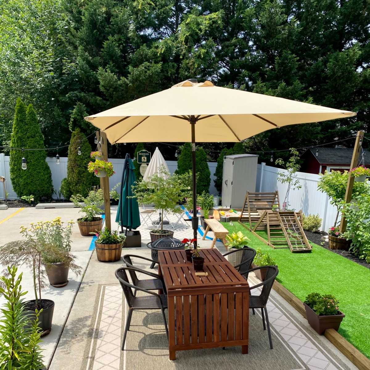 A patio area features a wooden table with chairs under a large umbrella, surrounded by potted plants and decorative greenery.