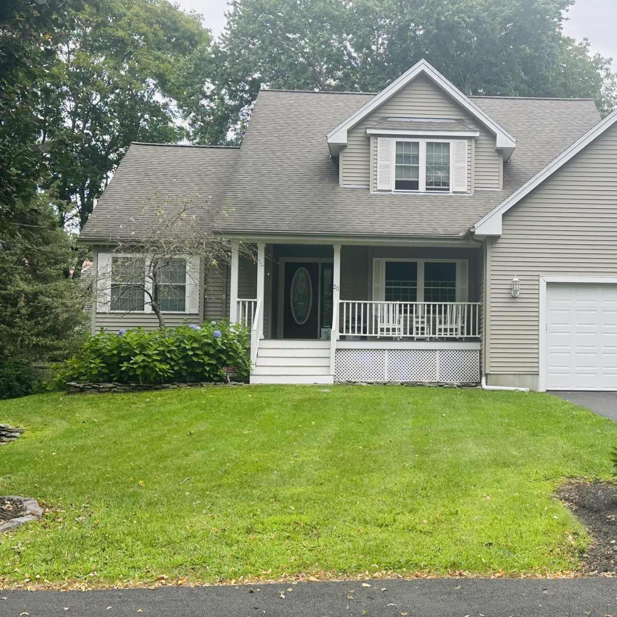 A two-story house with a peaked roof, front porch, and attached garage is situated on a green lawn surrounded by trees.