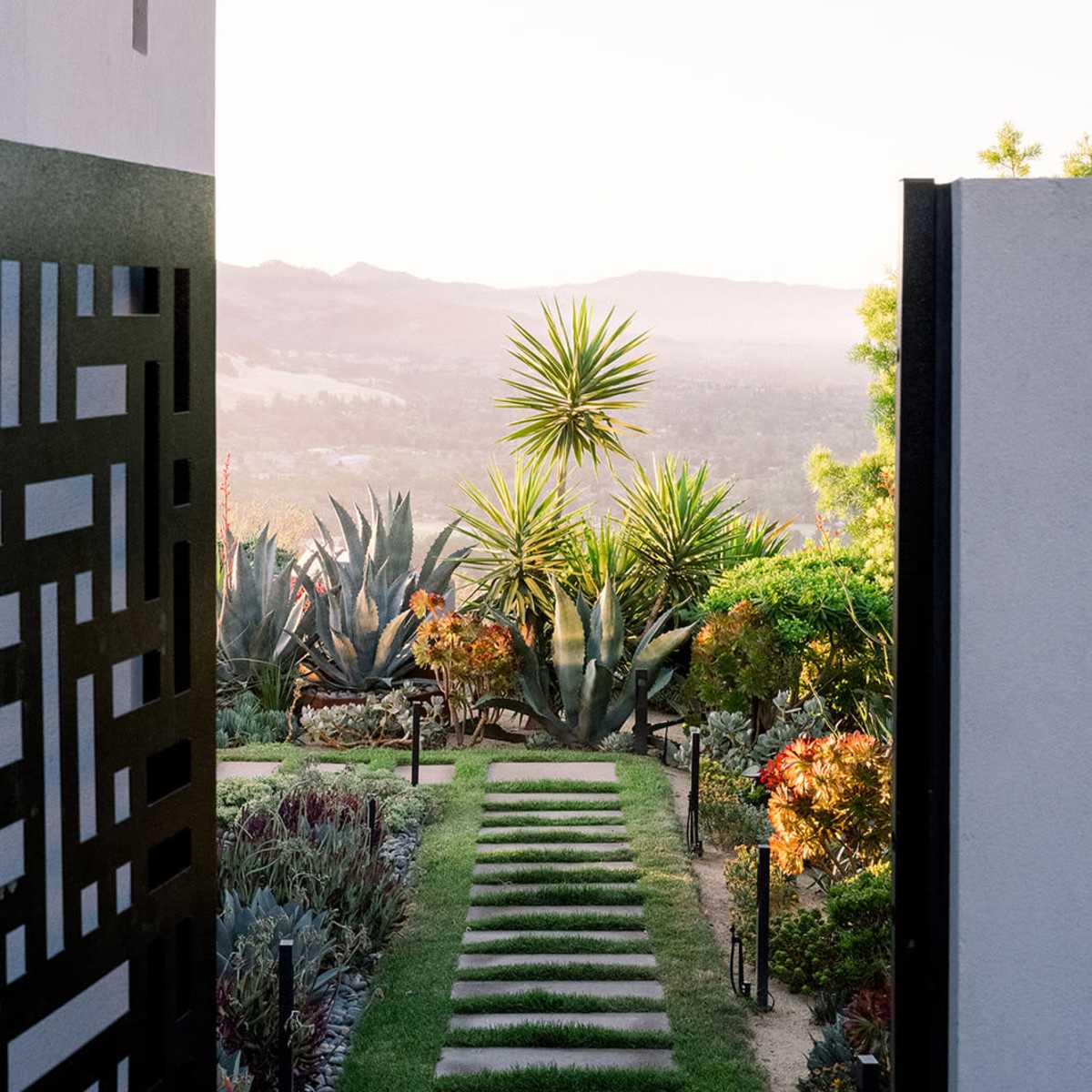 A stone pathway leads through a landscaped garden with various plants toward a scenic view of rolling hills at sunset.