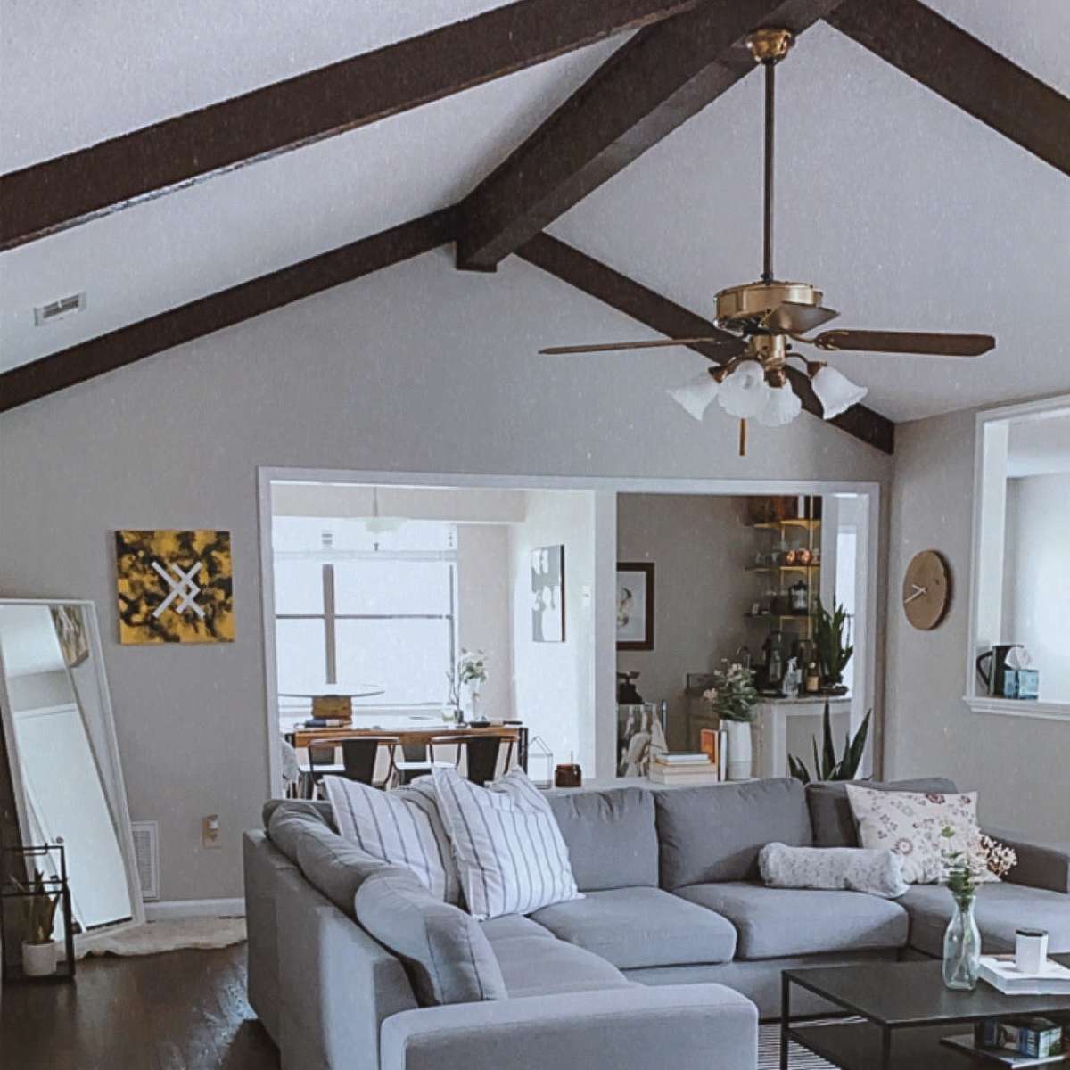 The living room features a gray sectional sofa, wooden beams on the ceiling, and a striped area rug on a hardwood floor.
