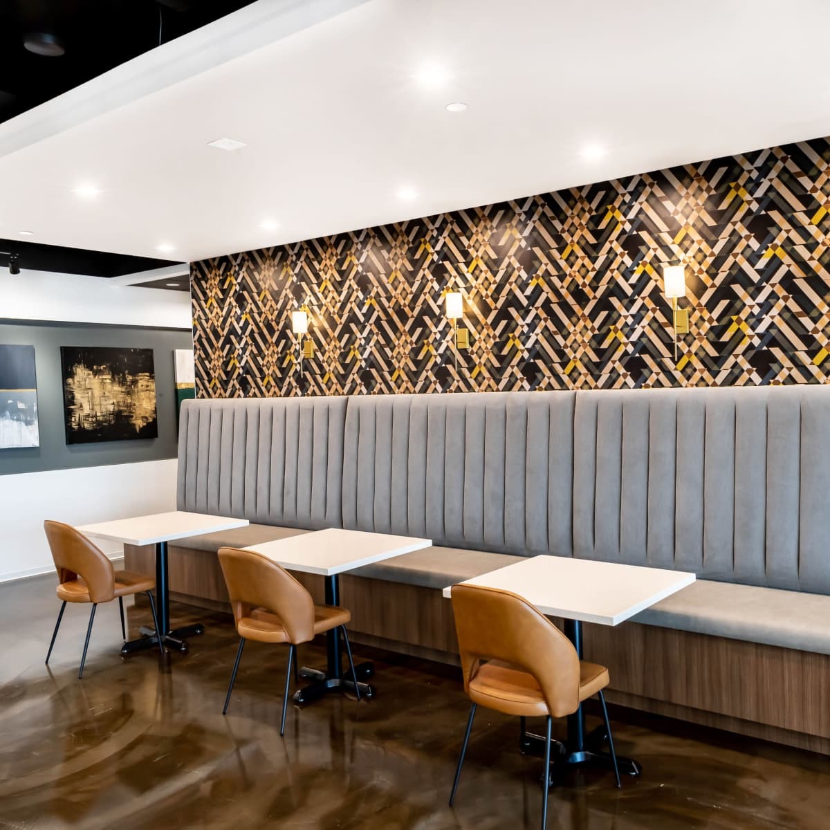 A modern dining area features a gray upholstered bench against a decorative wall and several small tables with brown chairs.