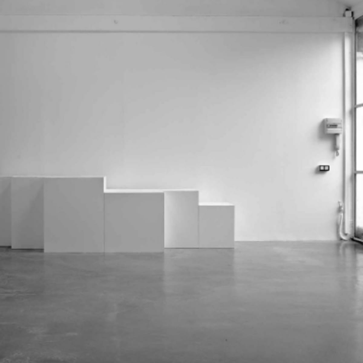 A minimalist gallery space featuring a series of white, stepped cuboid structures against a plain white wall.