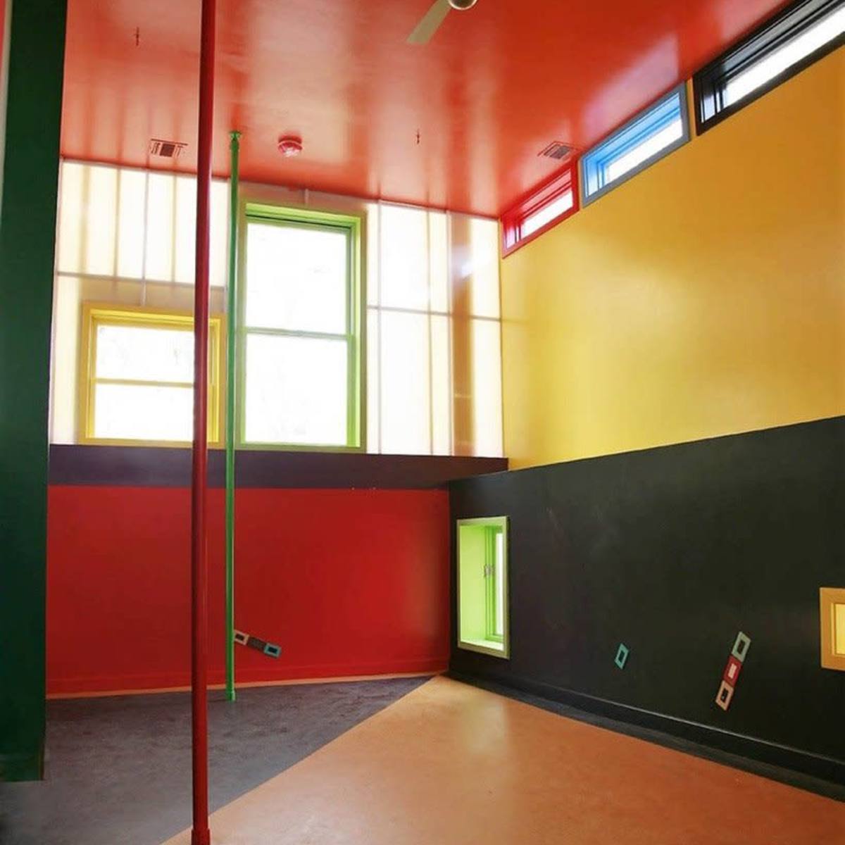 A brightly colored interior room featuring red, yellow, green, and black walls, with windows and a ceiling fan.