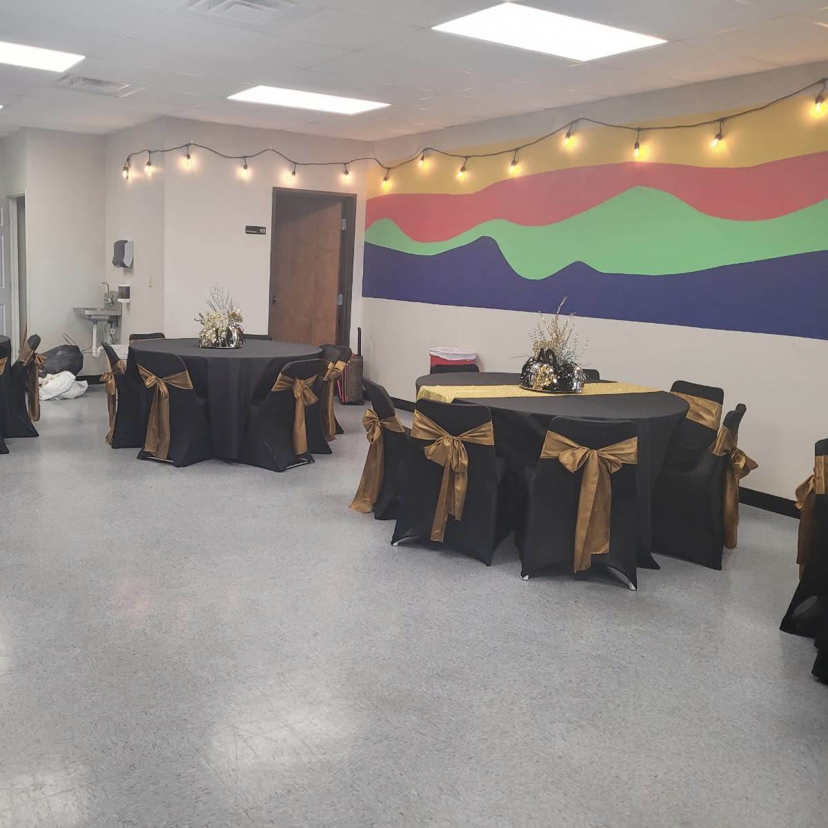 A room set up for an event, featuring several black tables adorned with gold bows and decorative centerpieces, illuminated by string lights on the walls.