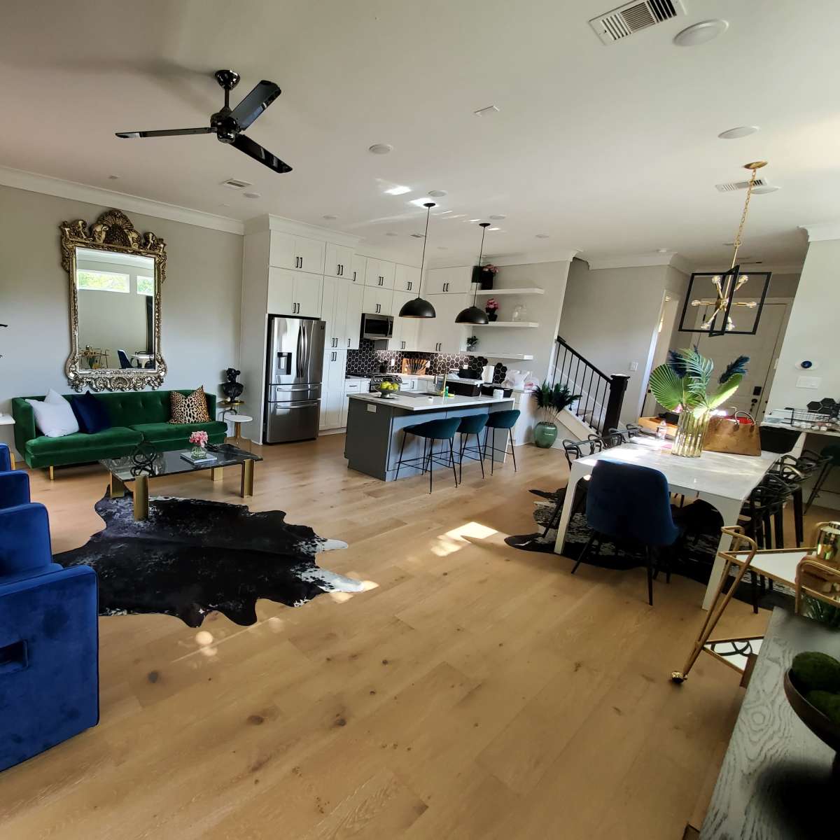 A spacious living area features a green velvet sofa, blue chairs, a large dining table, and an open kitchen with modern appliances.
