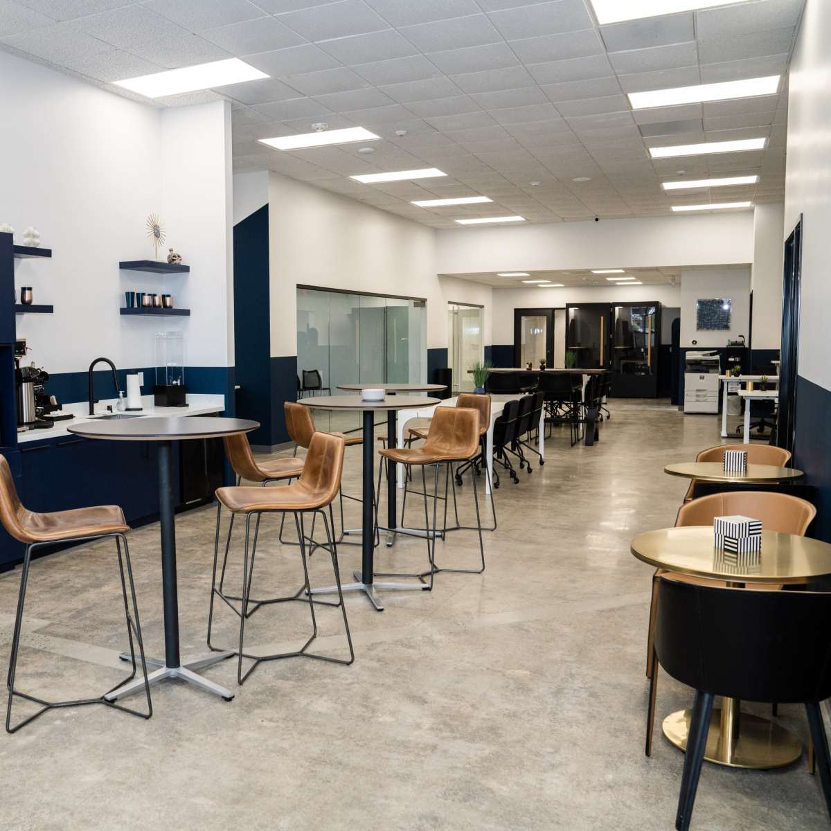 A modern office break area with high tables, bar stools, a fridge, and a coffee station.