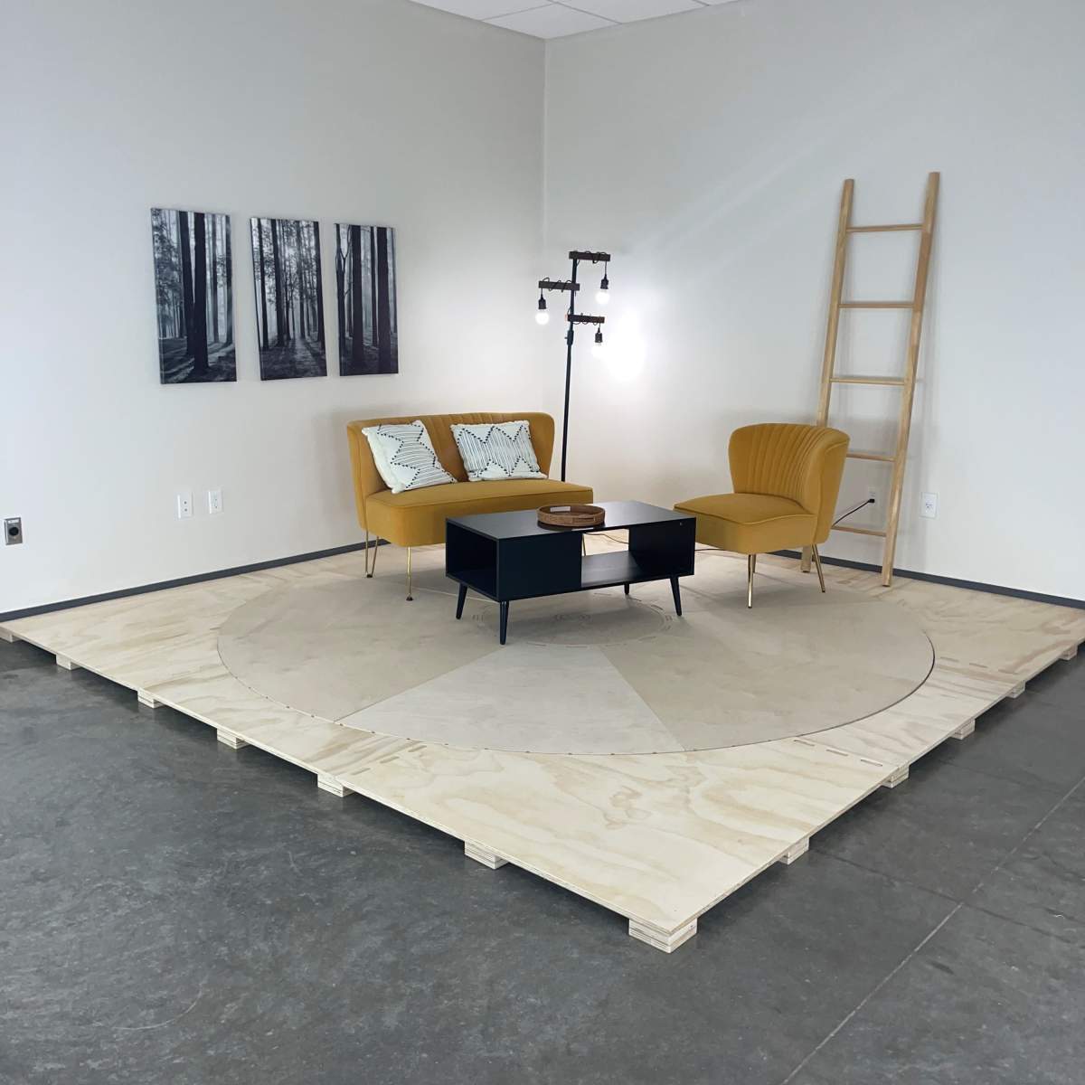 A minimalist room with two yellow armchairs, a black coffee table, a lamp, and a wooden ladder, all situated on a circular wooden platform surrounded by four framed pictures of trees.