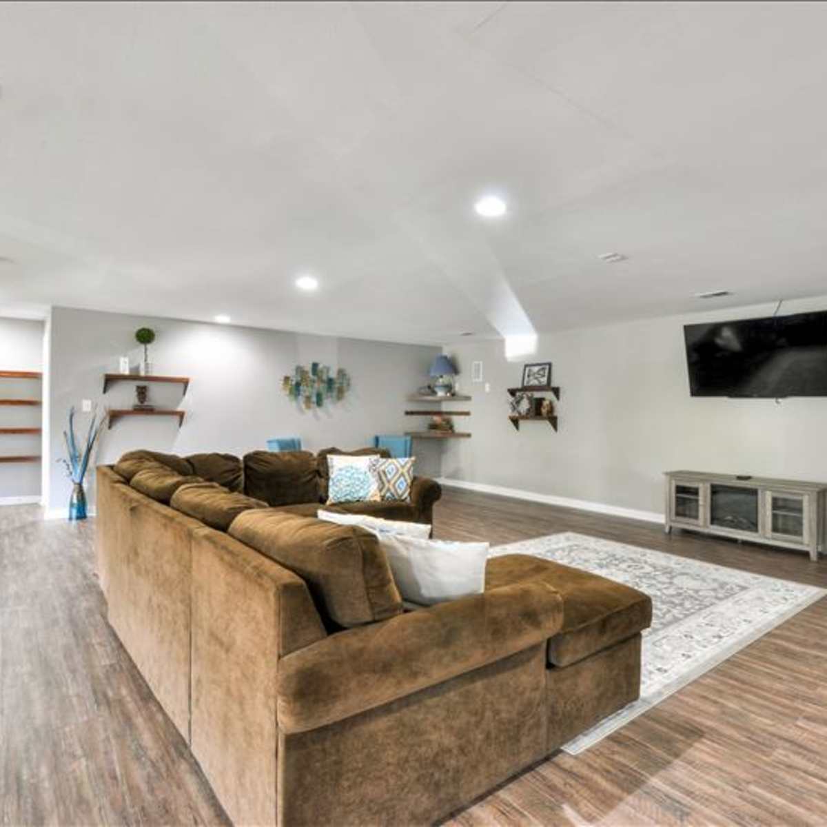 A spacious living room features a brown sectional sofa, a decorative area rug, and a wall-mounted television, with shelves and wall decor in the background.