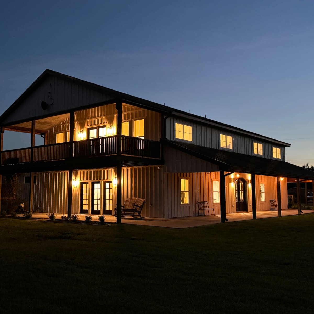 A two-story house with illuminated windows and porch lights stands on a green lawn at dusk.