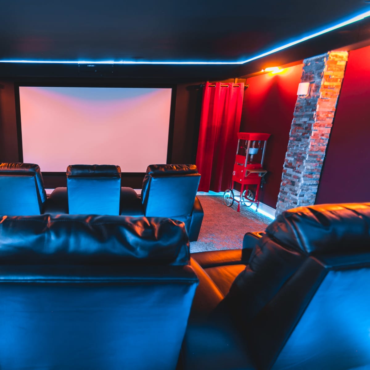 A home theater with black leather recliners facing a large screen, surrounded by red curtains and illuminated by blue accent lighting.