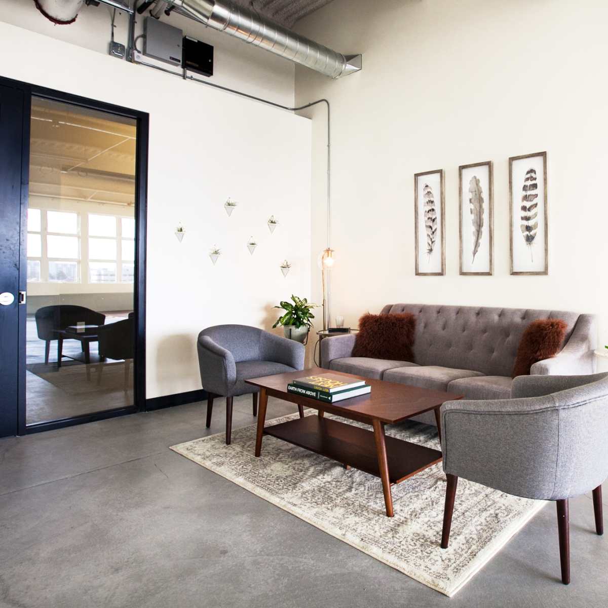 A modern, minimalistic lounge area features a gray sofa with decorative pillows, two matching armchairs, and a wooden coffee table, accented by framed artworks on the wall.
