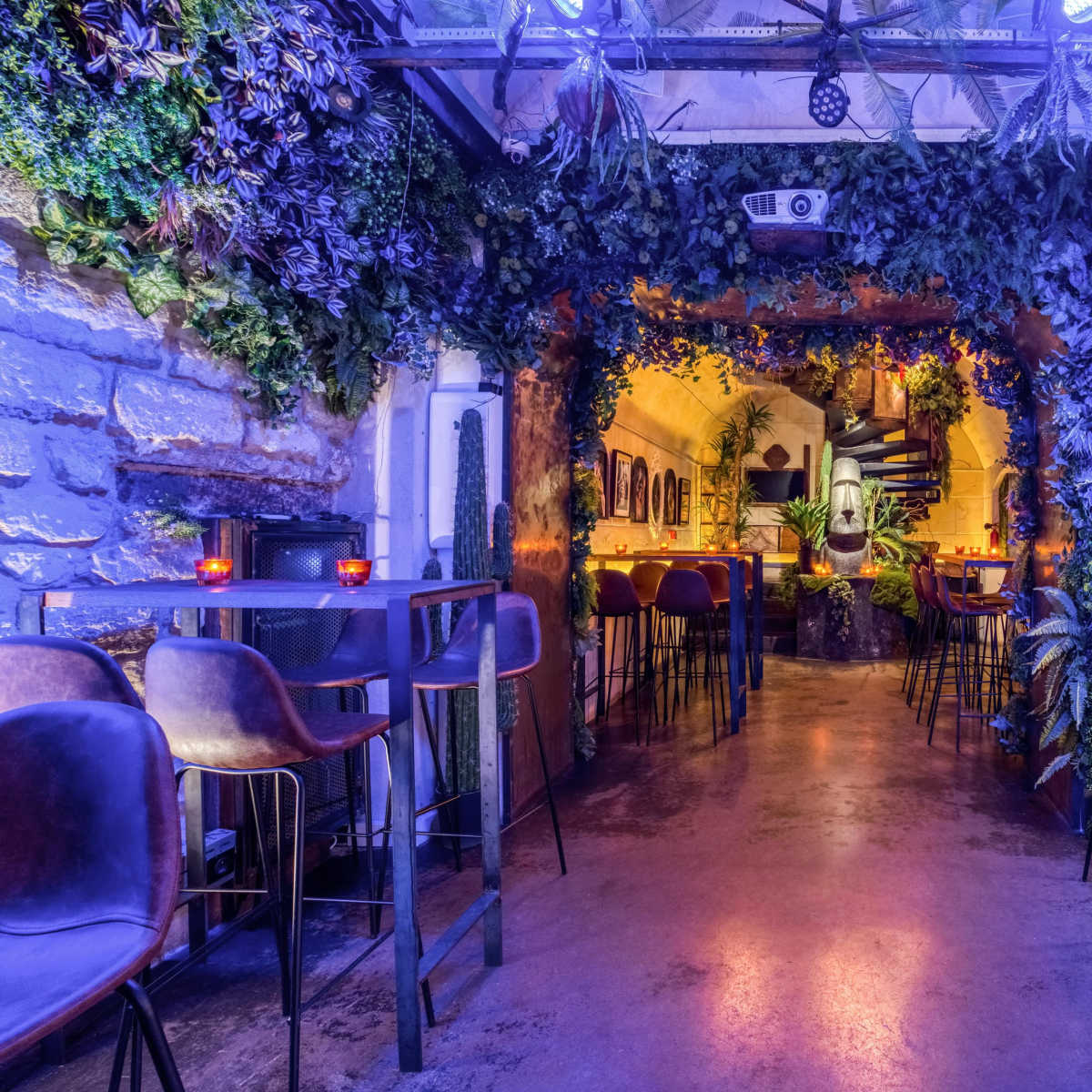 A dimly lit bar area with greenery-covered walls, small tables, and a staircase leading to another level.