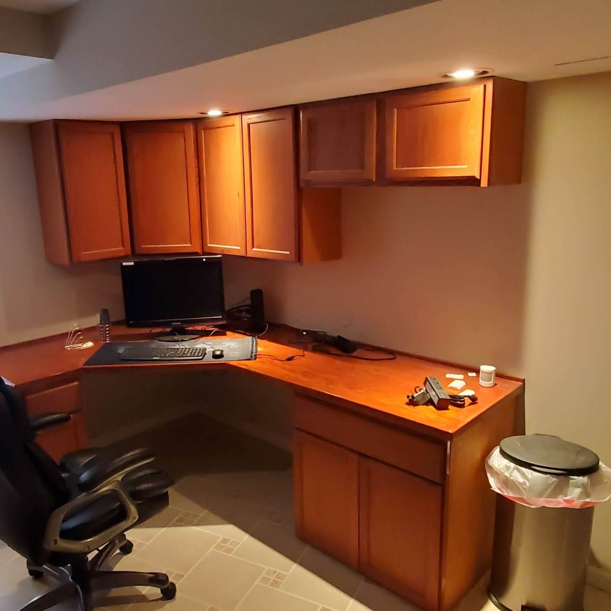 A wooden desk with a computer monitor, office chair, and trash can in a home office with a window featuring blinds.
