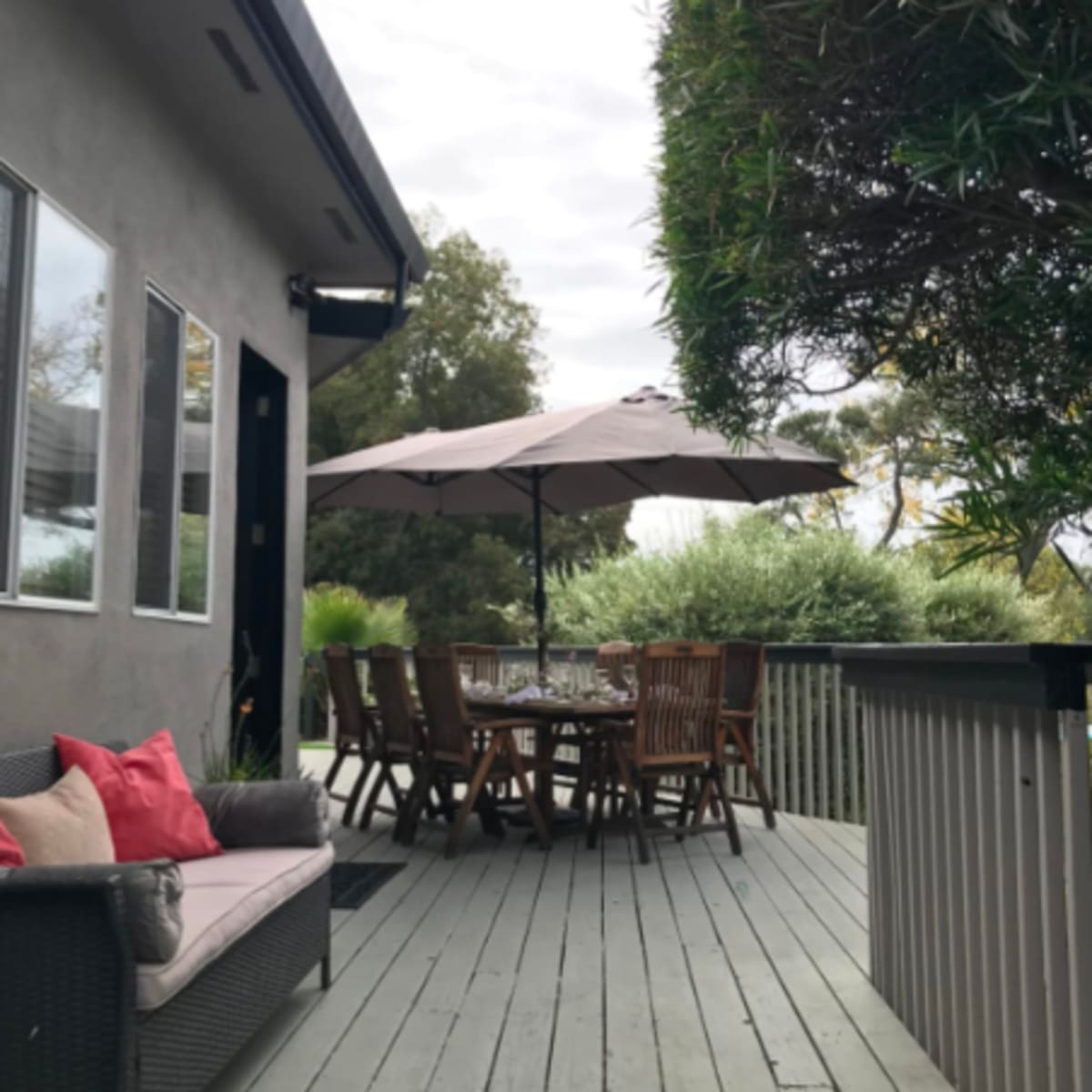 A wooden deck features a dining table with chairs, an umbrella, and a seating area, surrounded by greenery and a house in the background.