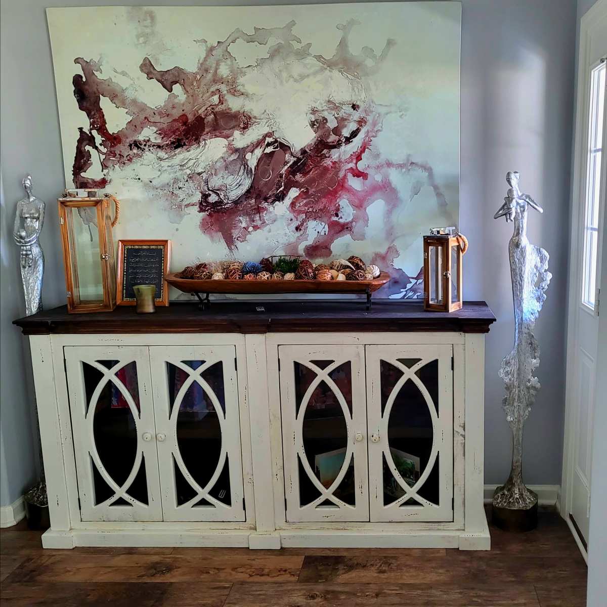 A white sideboard with glass doors displays decorative items, including candles and a large abstract painting, against a light gray wall.