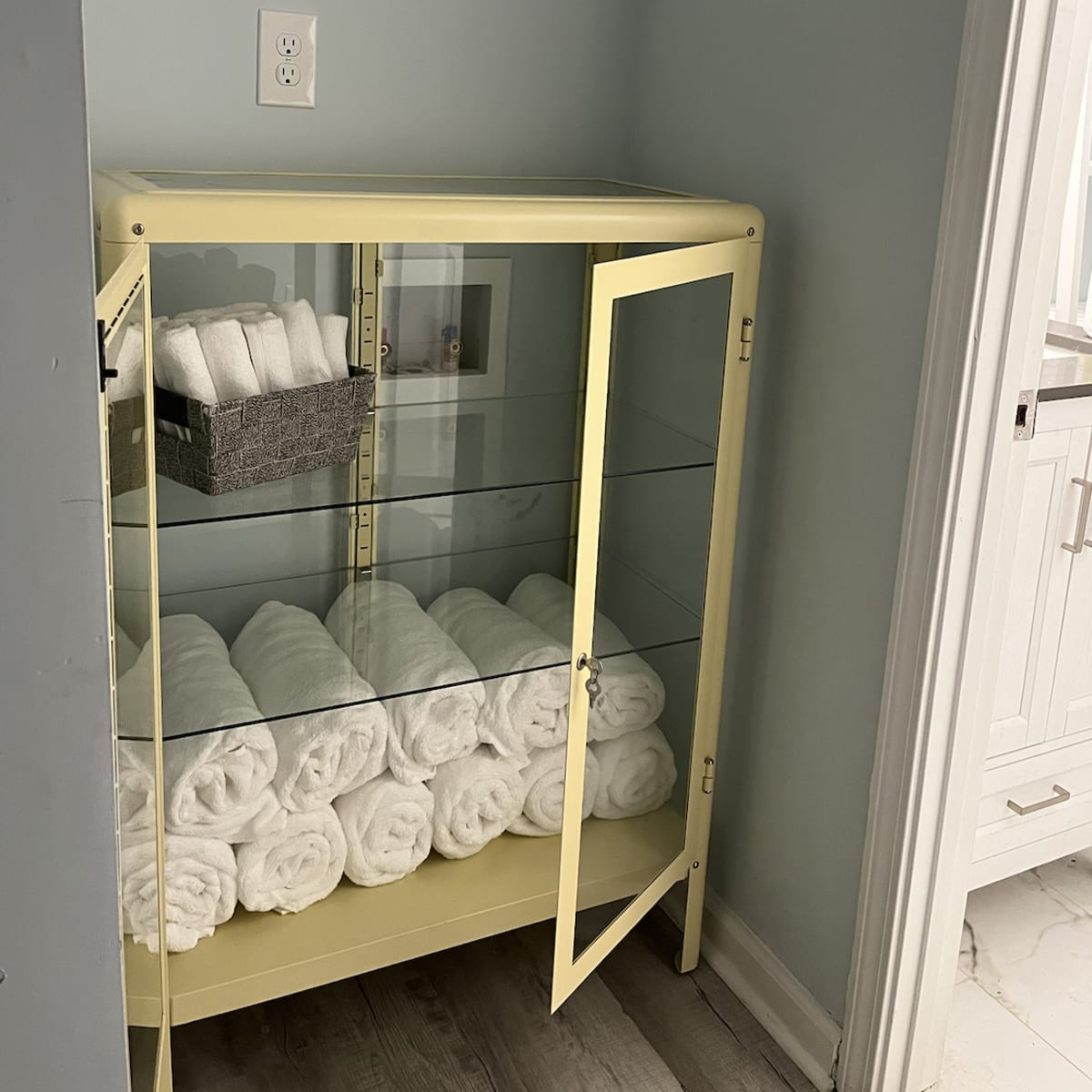 A yellow cabinet with glass doors displays neatly rolled white towels and a gray basket on a shelf.