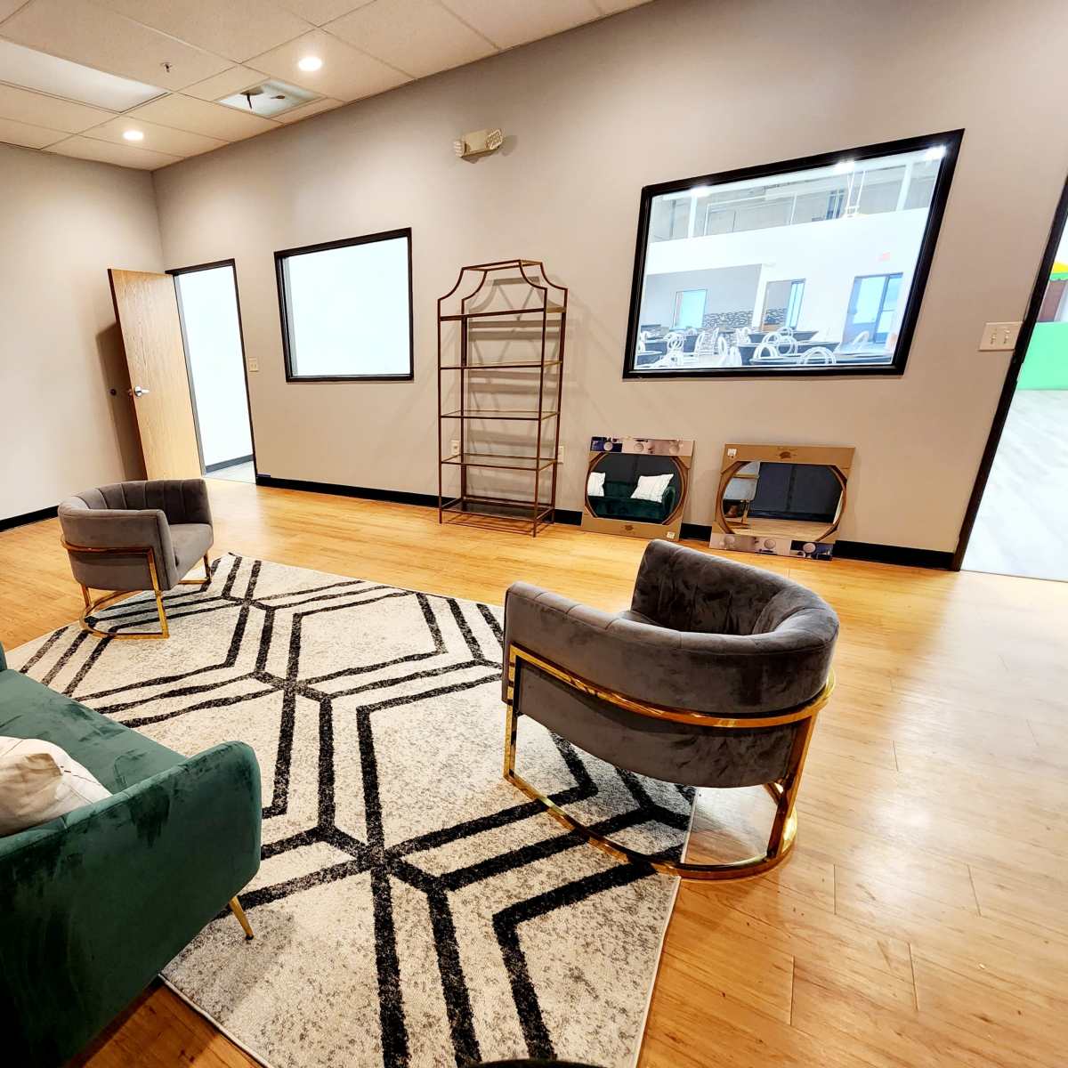 A minimalist lounge area with two velvet armchairs, a green sofa, and a geometric rug, along with a mirror and a large screen on the wall.