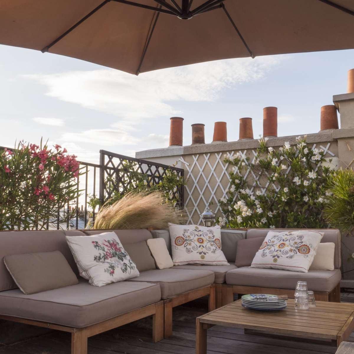 A rooftop terrace with a beige sofa set adorned with decorative pillows, a wooden coffee table, and potted plants under a large umbrella.