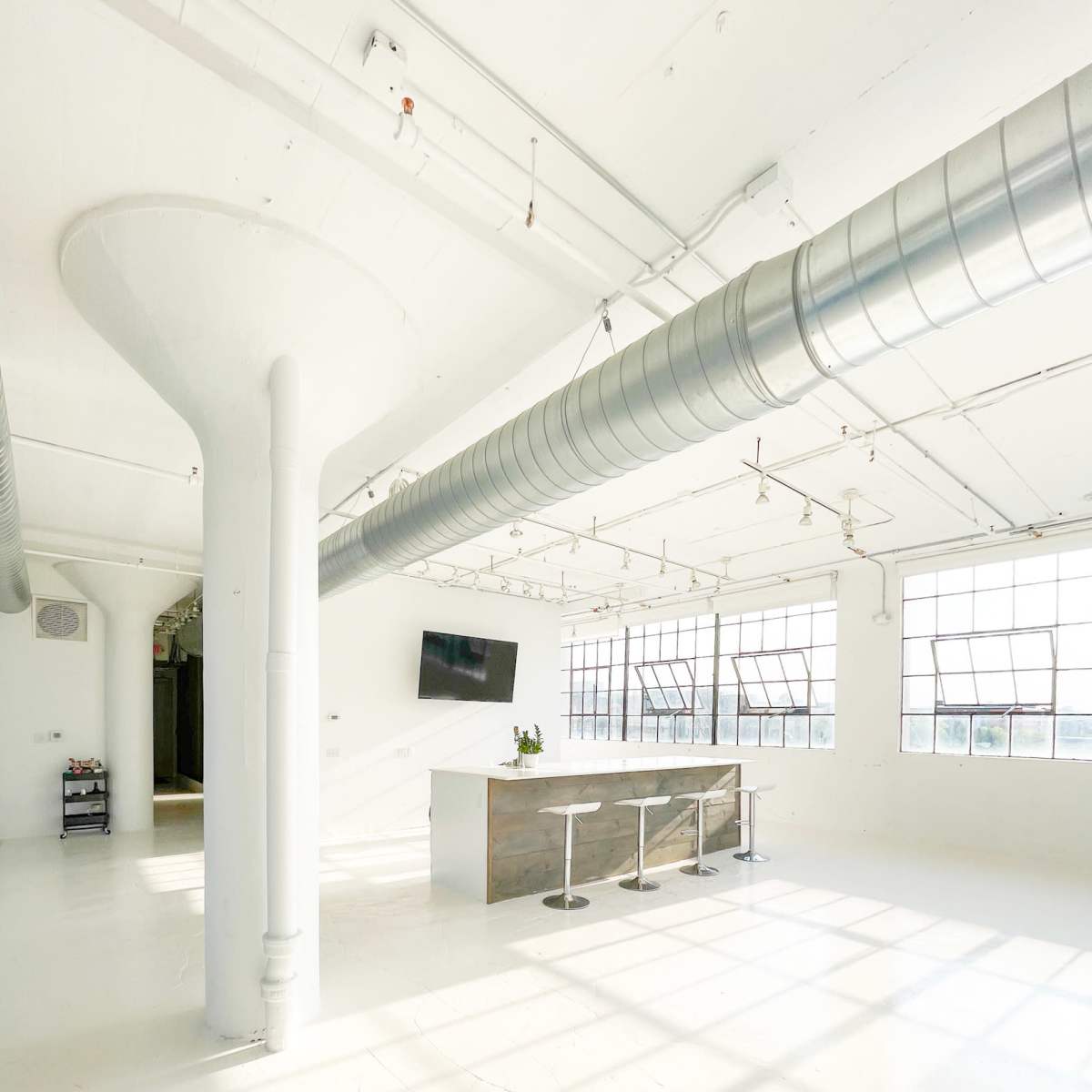 A bright, open space with white walls and floors, featuring a bar with three stools and large windows allowing natural light to fill the area.