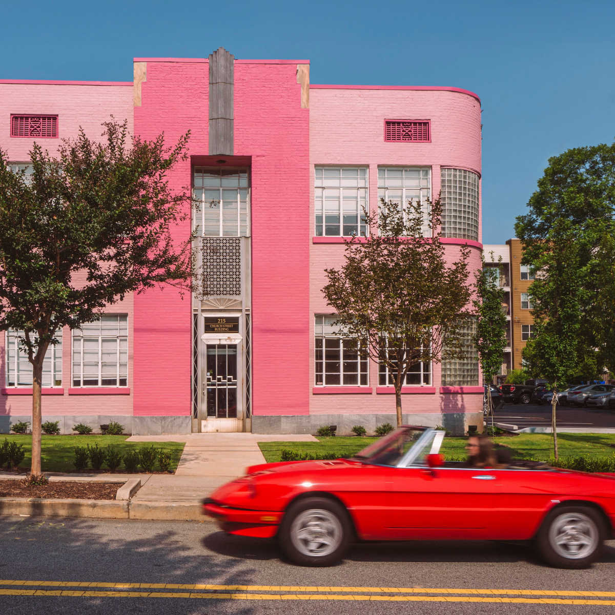 A bright pink building with glass brick accents stands alongside a tree-lined street as a red convertible drives past.