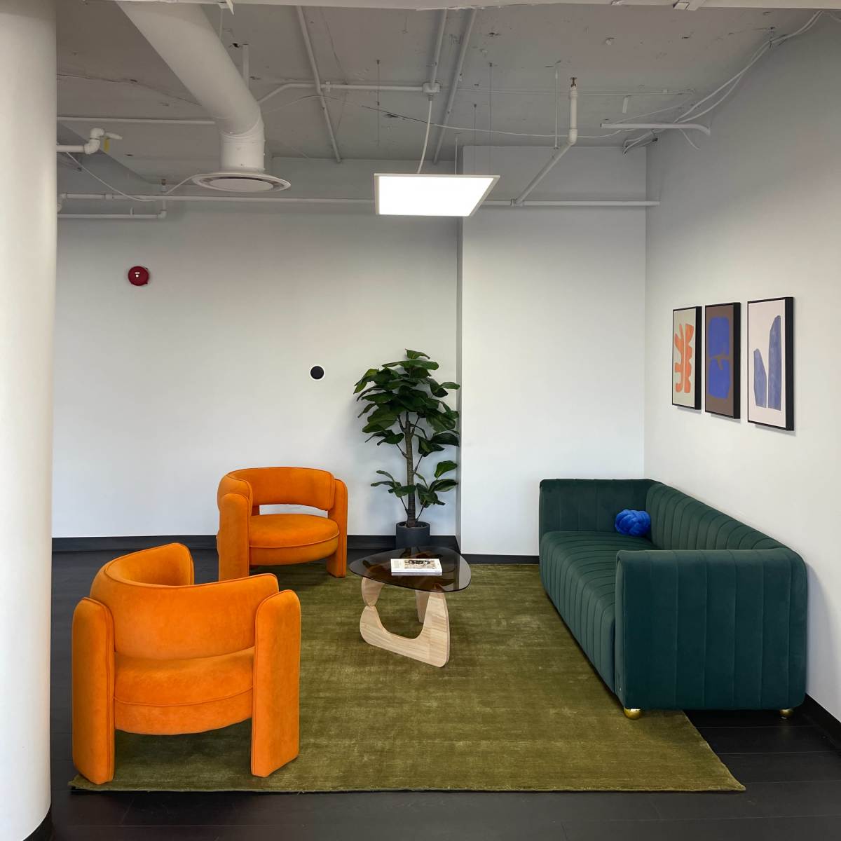 A modern seating area with two orange chairs, a green couch, a low coffee table, a rug, and artwork on the wall.