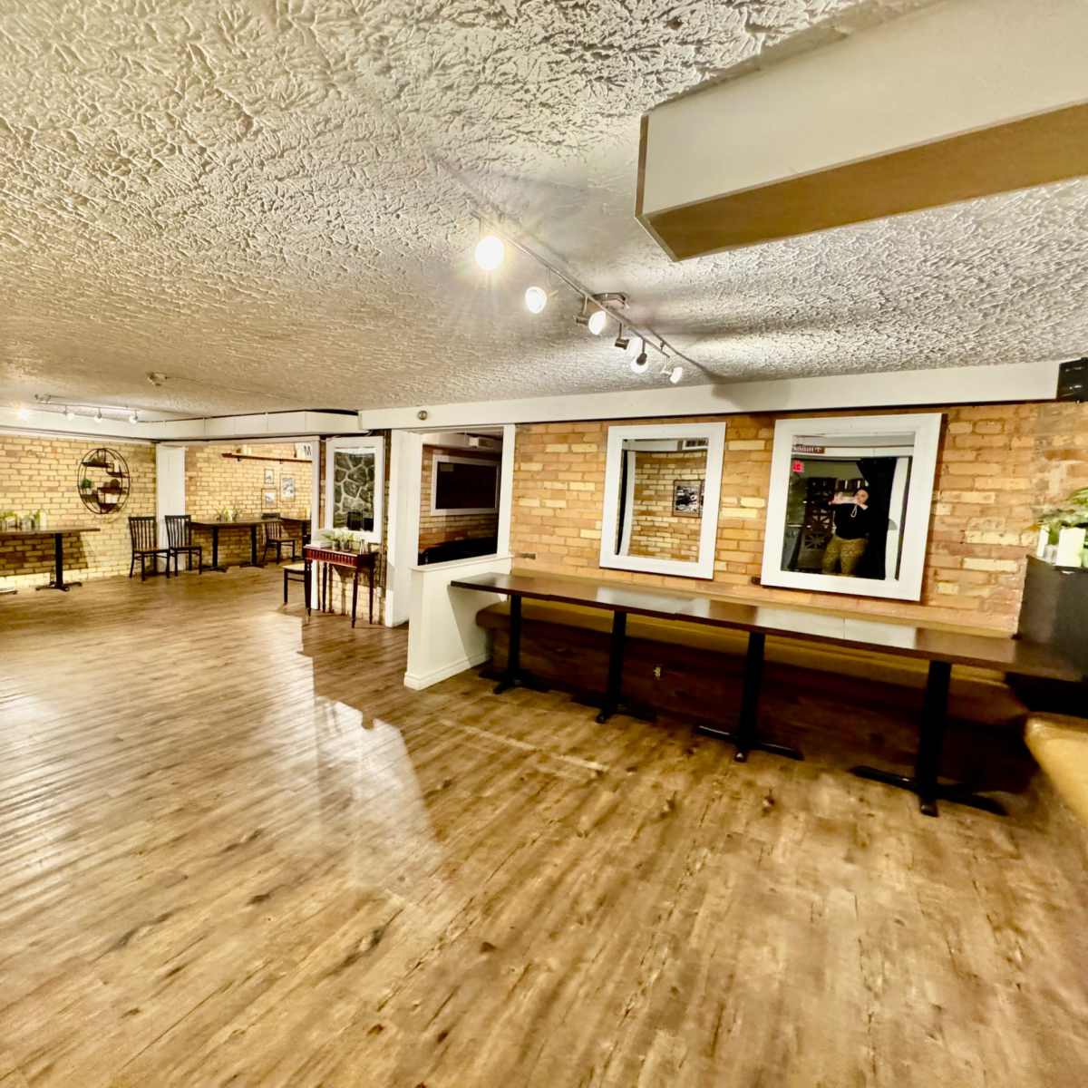 A spacious, well-lit room with wooden floors, brick walls, and several tables and chairs arranged around the area.
