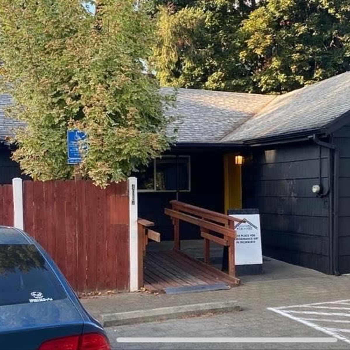 A black wooden building with multiple windows and a ramp leads to the entrance, surrounded by trees and parked cars.