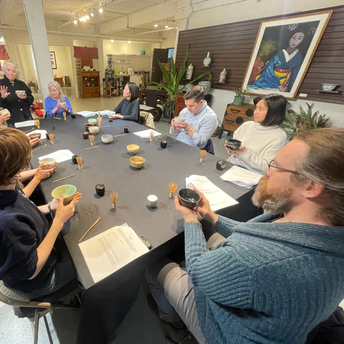 A group of people sits around a large table in a workshop, holding bowls and attentively listening to a speaker.