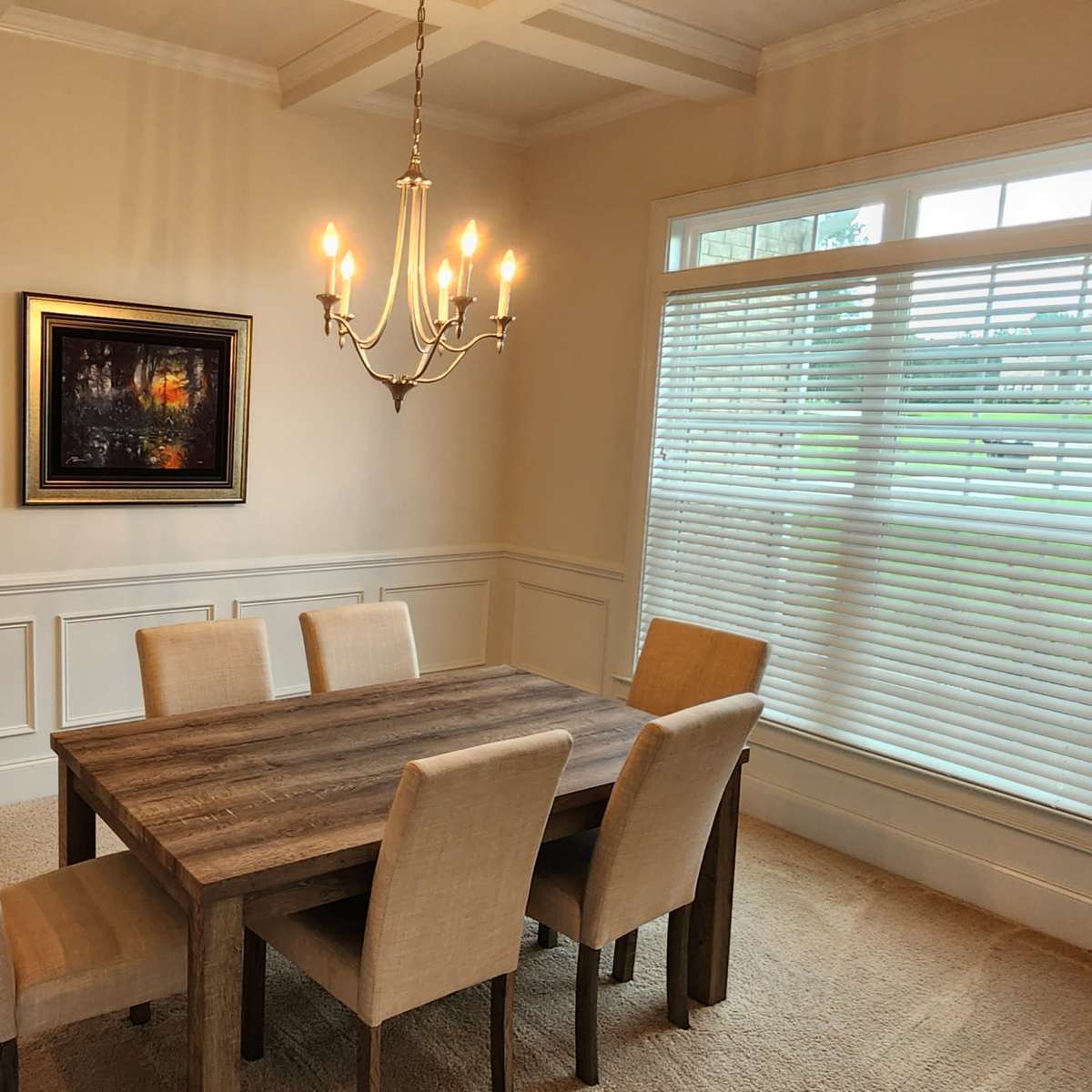 A dining room features a wooden table surrounded by six chairs, with a chandelier overhead and a large window providing natural light.