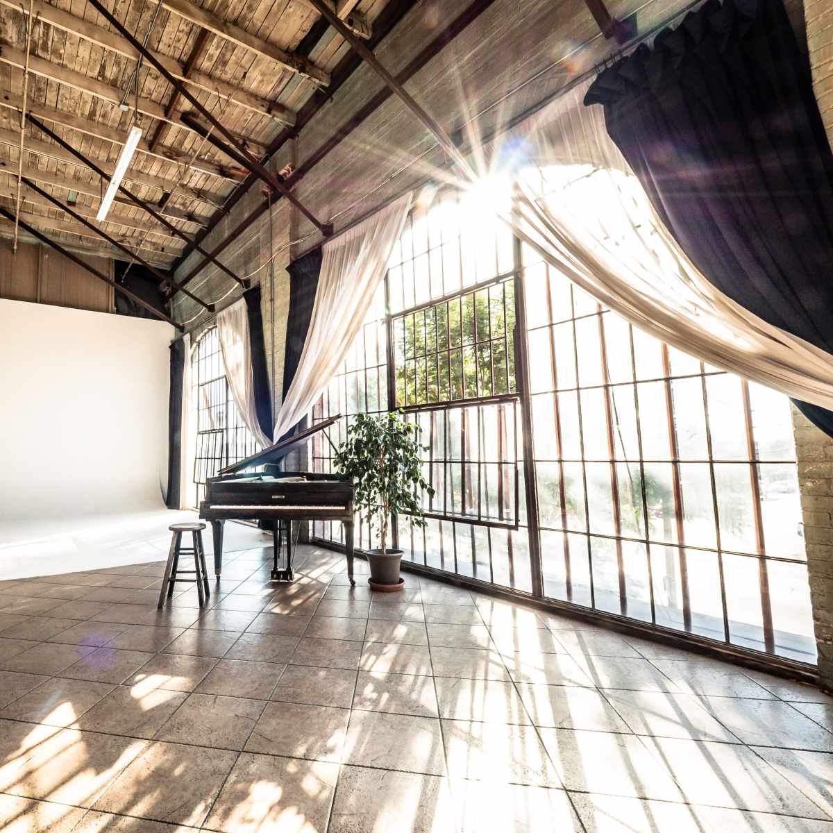 An open, sunlit space featuring large windows, a grand piano, and a stool, accented by draped fabric and a potted plant.