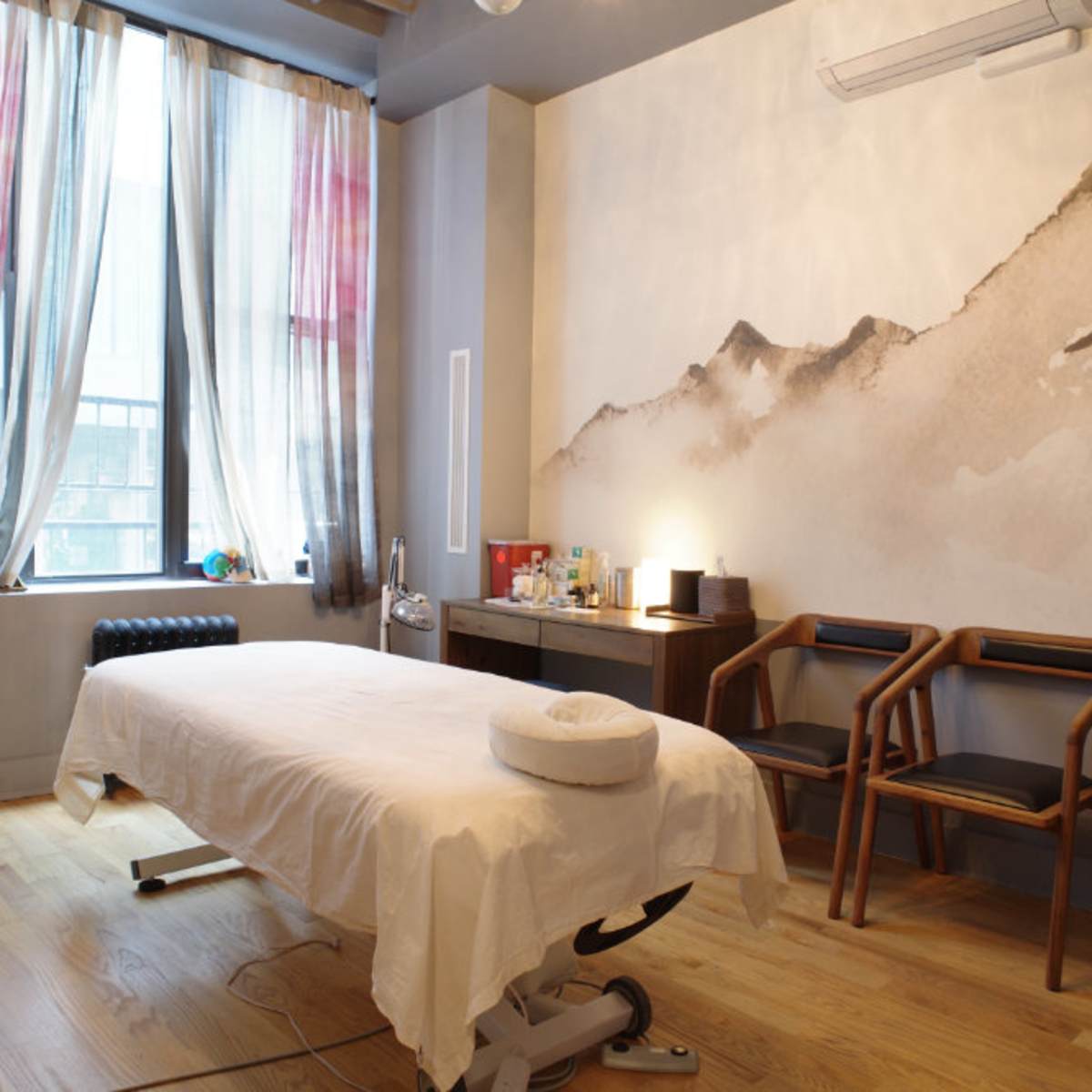 A treatment room with a massage table, wooden chairs, and a large mural of mountains on the wall.