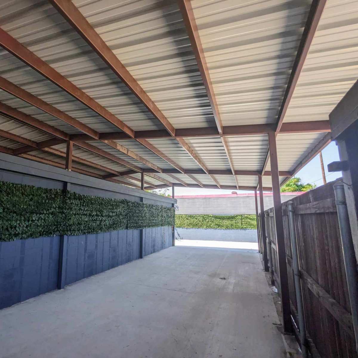 A covered pathway with metal beams and a greenery-covered wall on one side.