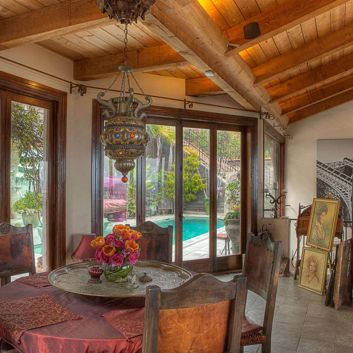 A sunlit dining area with a round table, ornate light fixture, and views of a pool and garden through large windows.