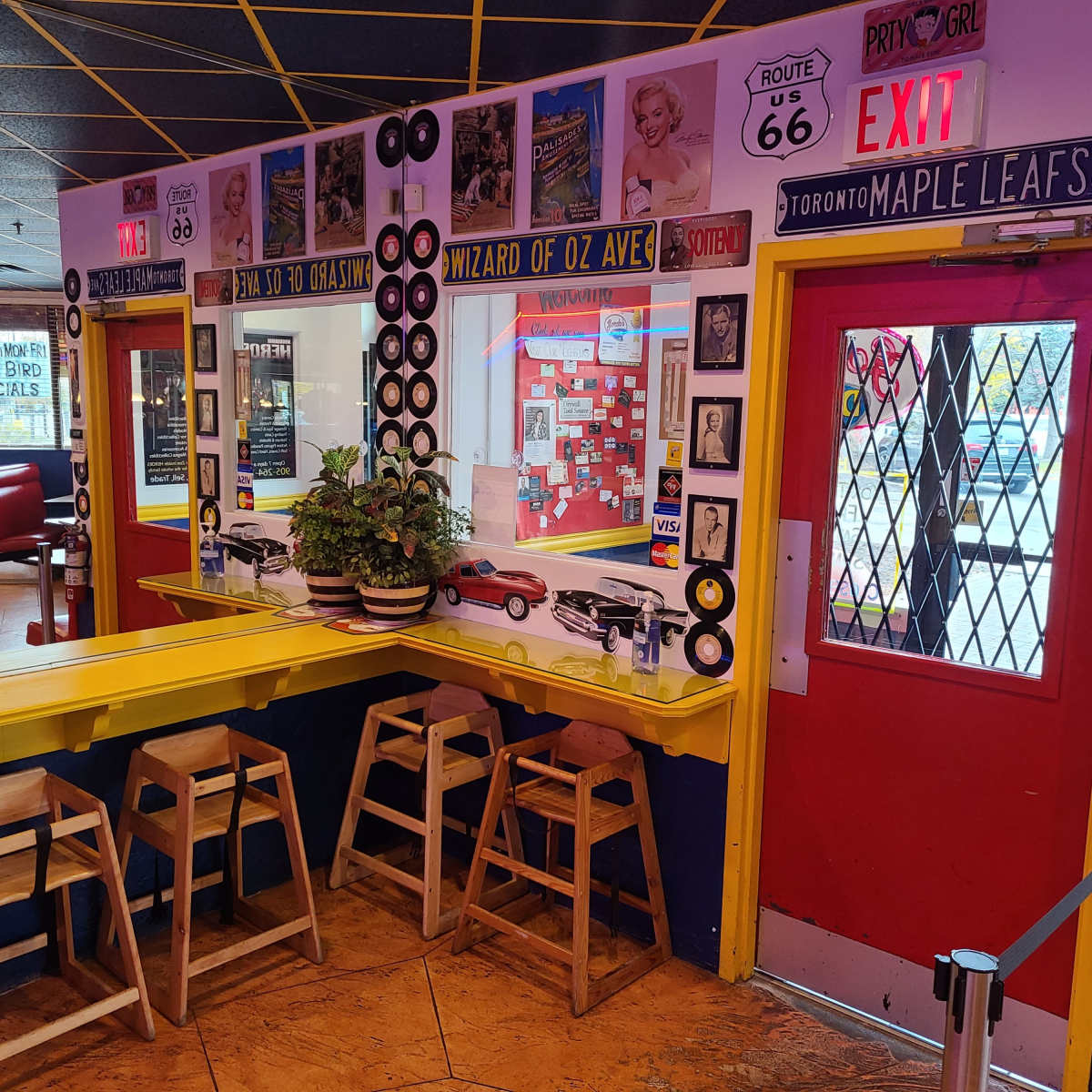 A brightly colored diner interior featuring a yellow bar with wooden stools, decorative vinyl records on the walls, and a red exit door next to various memorabilia.