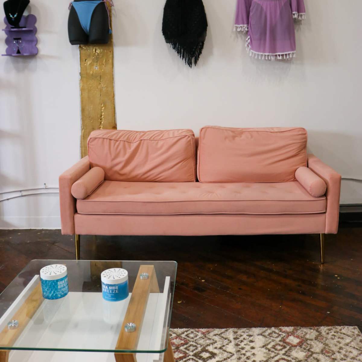 A pink couch in front of a glass coffee table, with various garments displayed on mannequins against a wall.