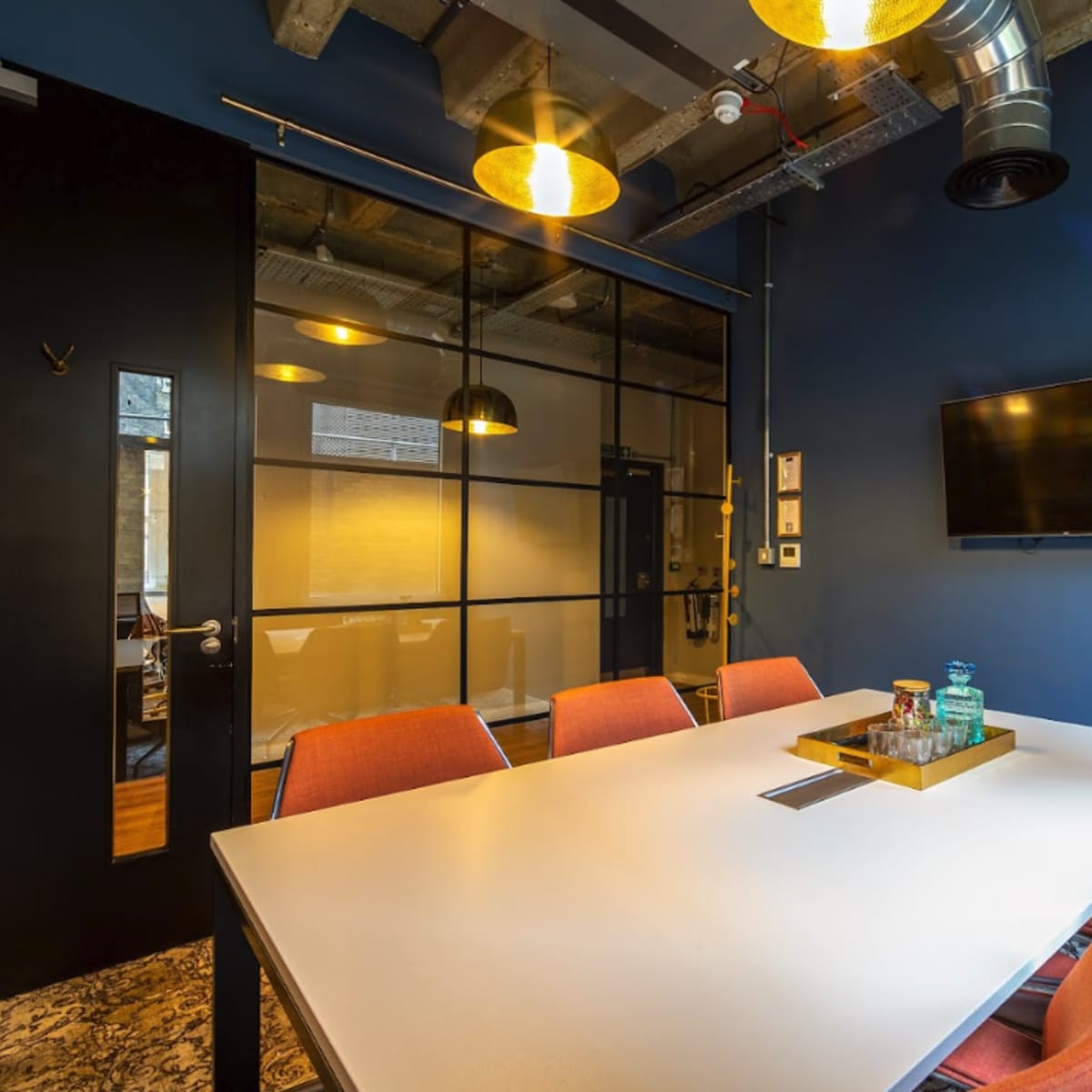 A modern conference room with a white table, orange chairs, a glass wall, and a wall-mounted television.